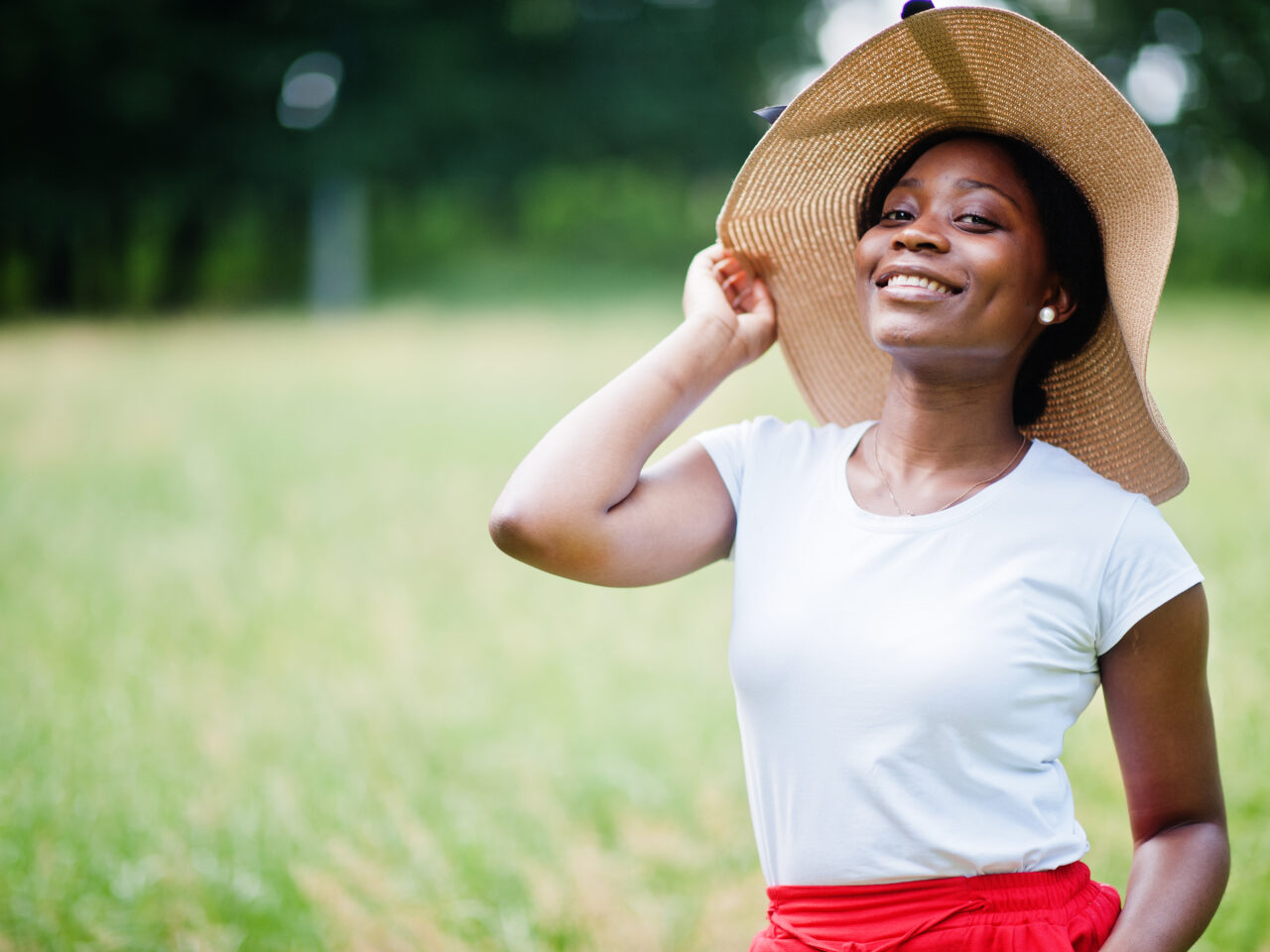 Portrait of gorgeous african american woman 20s wear in summer hat, red pants and white t-shirt, posing at green grass in park.